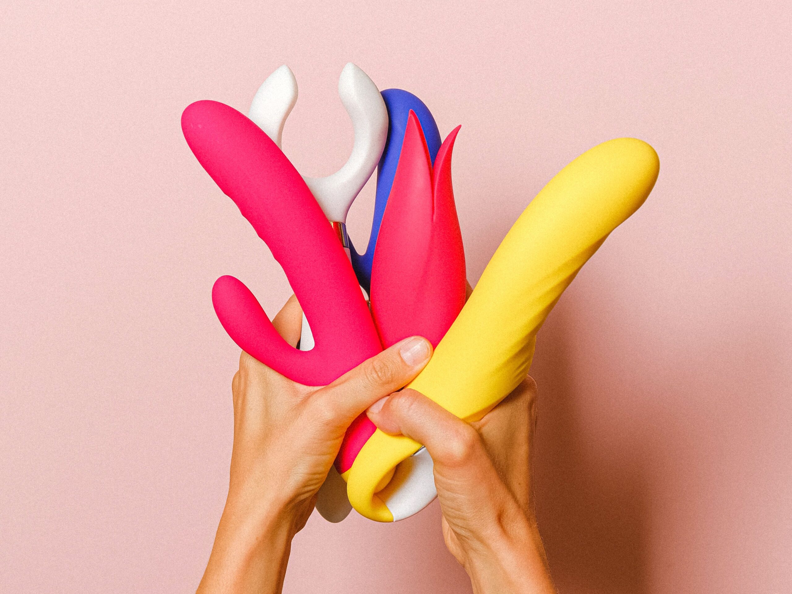 Hands holding vibrant silicone vibrators, showcasing diverse designs against a pink backdrop.