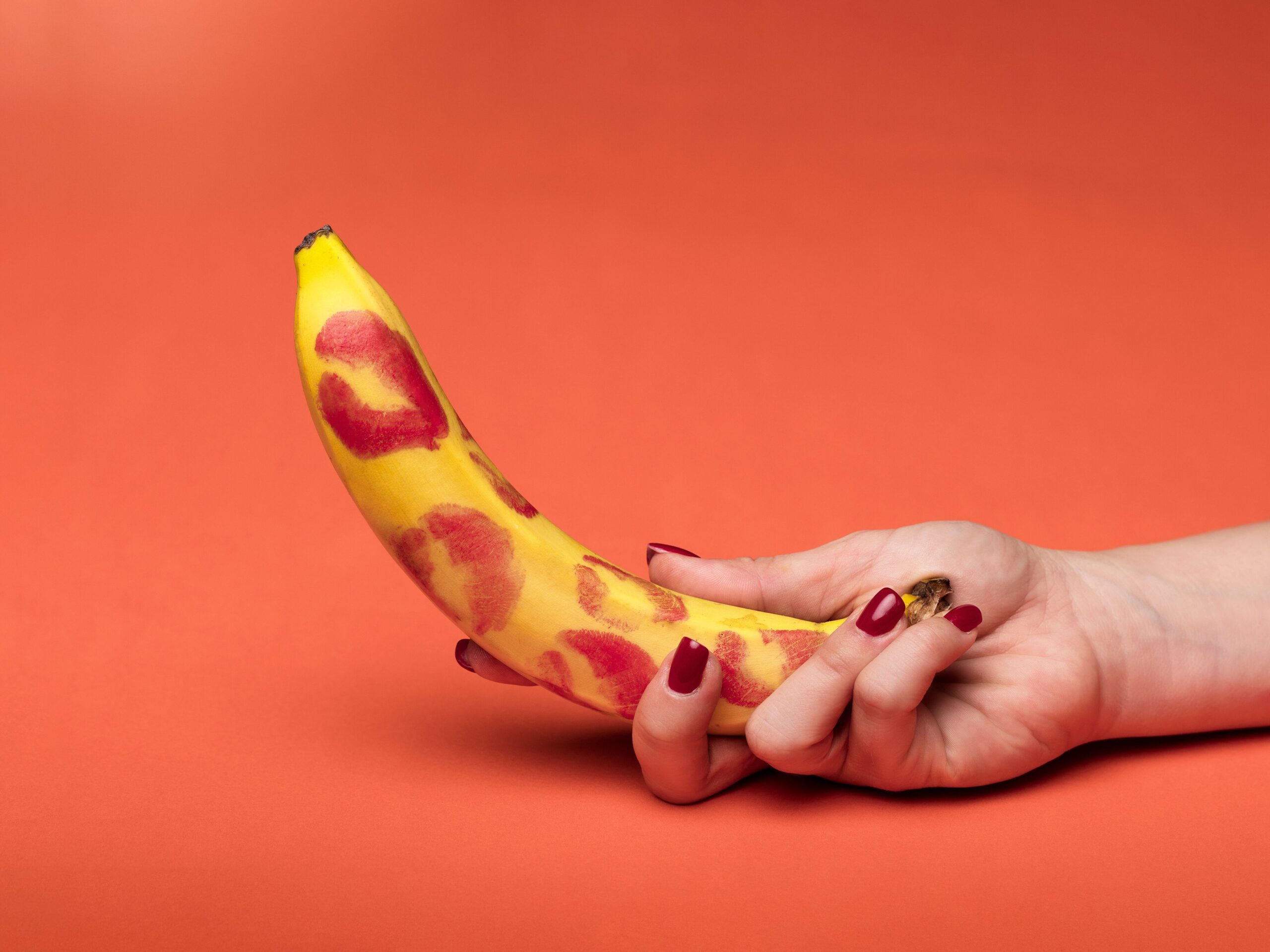 A feminine hand holds a banana covered in lipstick marks on a vibrant red background.