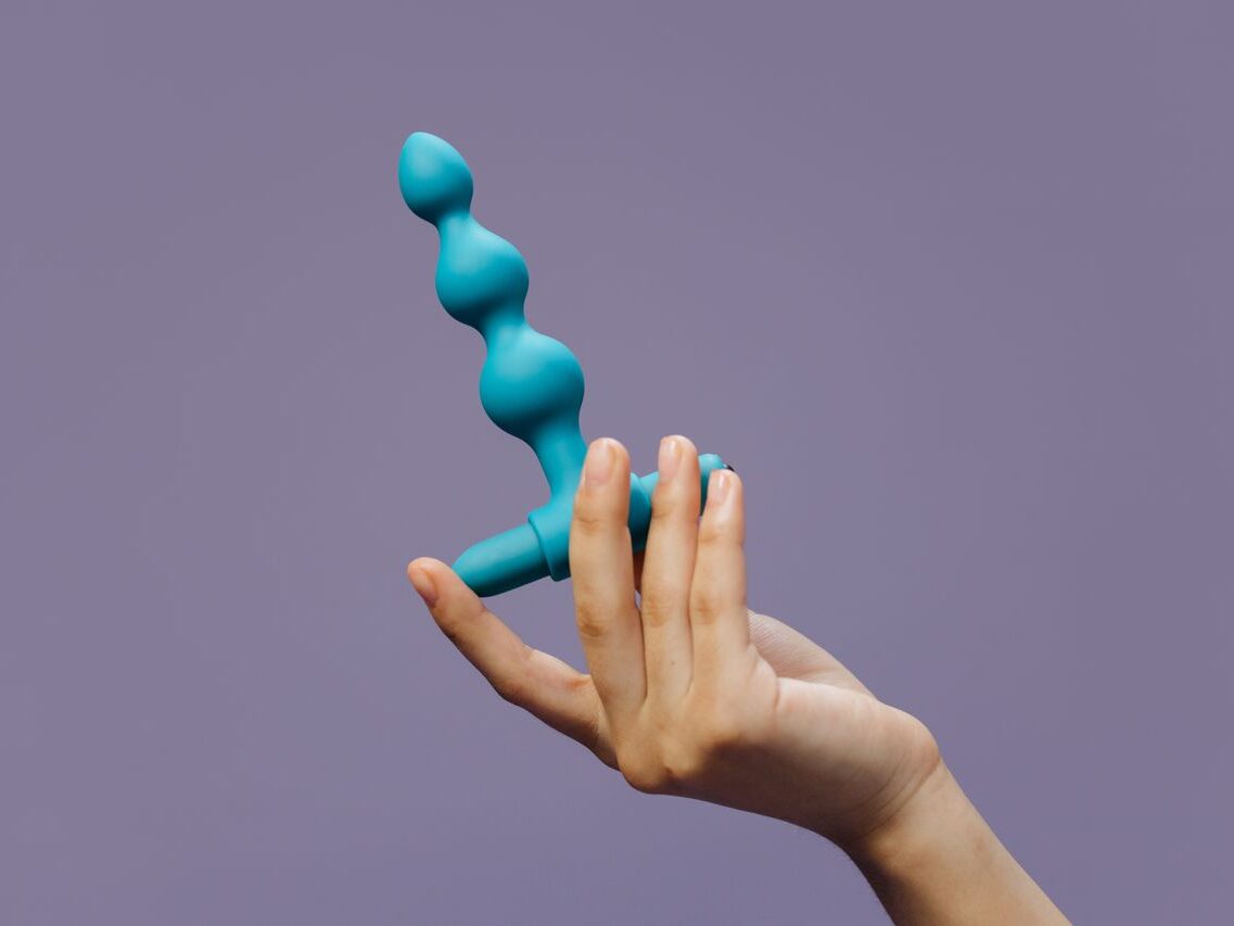 Close-up of a hand holding blue silicone anal beads against a purple background highlighting sensuality and self-love.