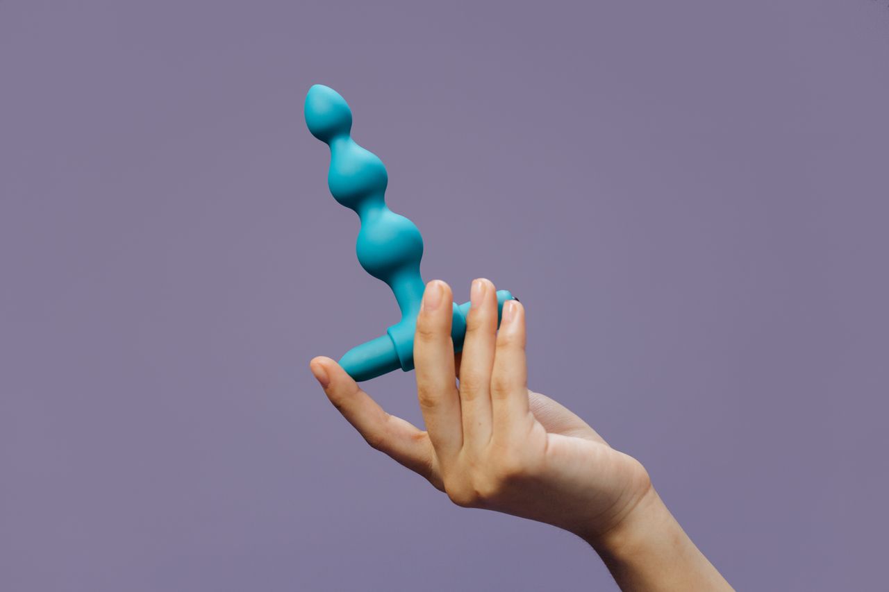 Close-up of a hand holding blue silicone anal beads against a purple background highlighting sensuality and self-love.